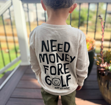 A child viewed from behind wearing a light beige long-sleeve shirt with bold black text reading 'NEED MONEY FORE GOLF' and olive green pants, standing on a wooden deck outdoors.