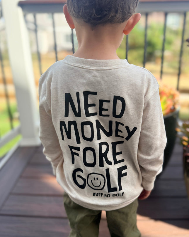 A child viewed from behind wearing a light beige long-sleeve shirt with bold black text reading 'NEED MONEY FORE GOLF' and olive green pants, standing on a wooden deck outdoors.