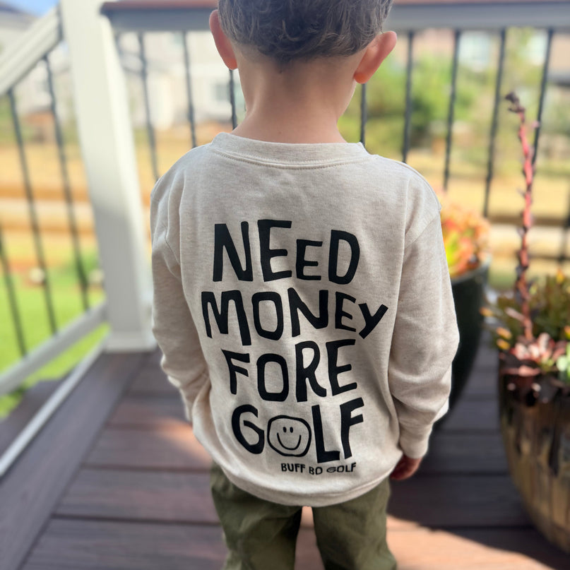 A child viewed from behind wearing a light beige long-sleeve shirt with bold black text reading 'NEED MONEY FORE GOLF' and olive green pants, standing on a wooden deck outdoors.