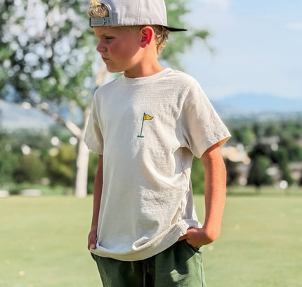 Young boy wearing a white t-shirt with a logo and green shorts, standing outdoors.