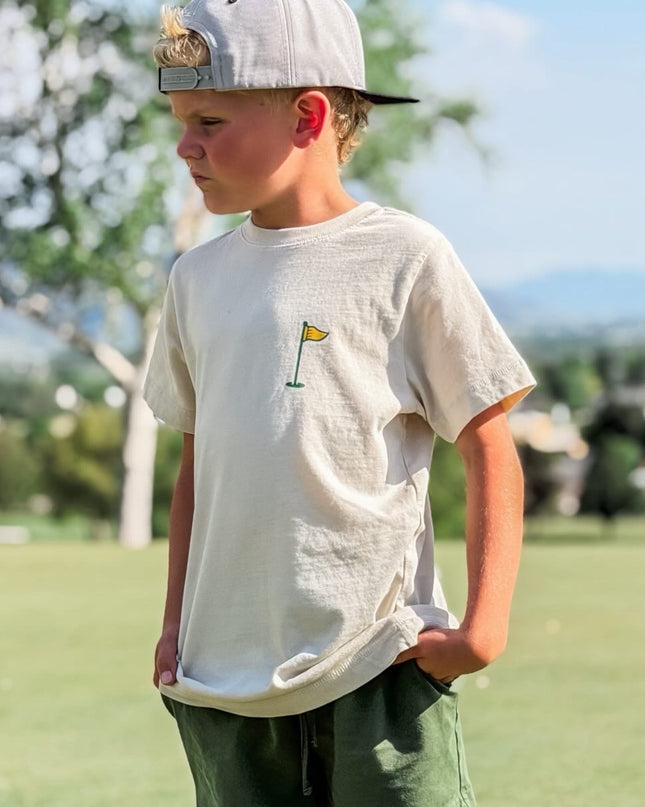 Young boy wearing a white t-shirt with a logo and green shorts, standing outdoors.