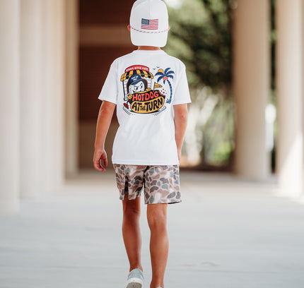 A child walking away in a covered walkway, wearing a white t-shirt with a large colorful 'SUNSHINE CANYON' graphic on the back, camouflage shorts, and a white cap.