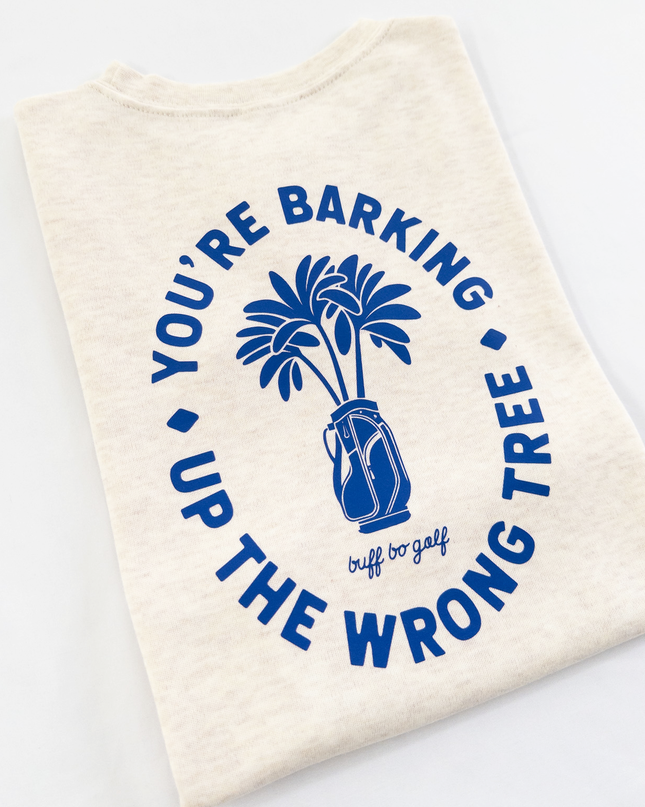 Beige kids golf t-shirt featuring bold blue graphic with palm trees in a golf bag and playful text reading 'You're Barking Up The Wrong Tree,' from Buff Bo Golf apparel collection, displayed on a light background.
