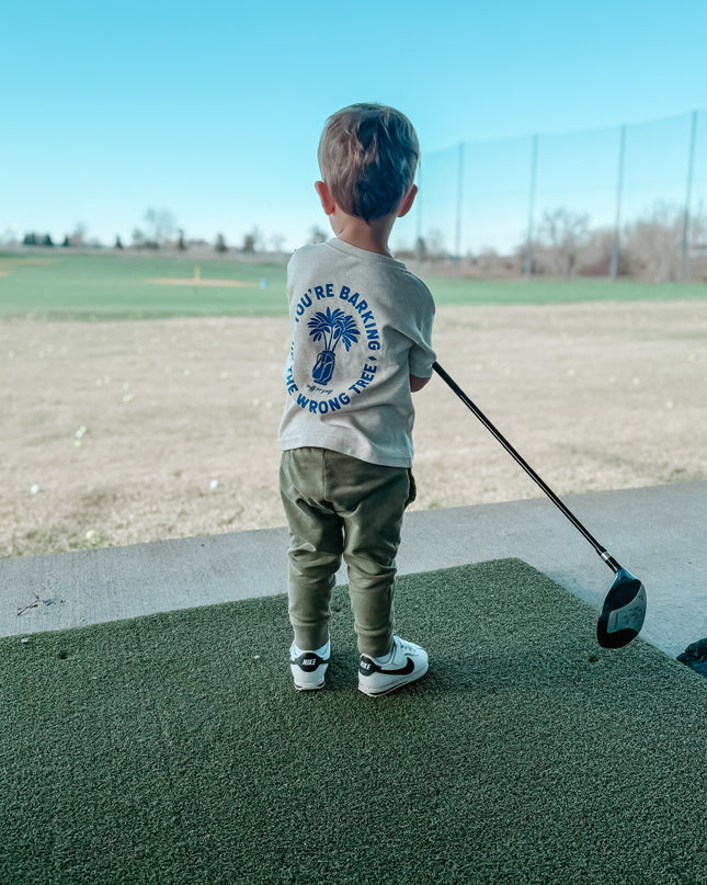 Young child holding a golf driver while wearing a beige t-shirt with blue graphic reading 'You’re Barking Up The Wrong Tree,' standing on a driving range with an open golf course view, showcasing kids golf apparel in action.