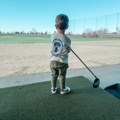Young child holding a golf driver while wearing a beige t-shirt with blue graphic reading 'You’re Barking Up The Wrong Tree,' standing on a driving range with an open golf course view, showcasing kids golf apparel in action.