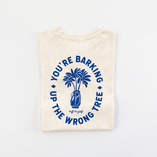 Beige kids golf t-shirt featuring bold blue graphic with palm trees in a golf bag and playful text reading 'You're Barking Up The Wrong Tree,' from Buff Bo Golf apparel collection, displayed on a light background.