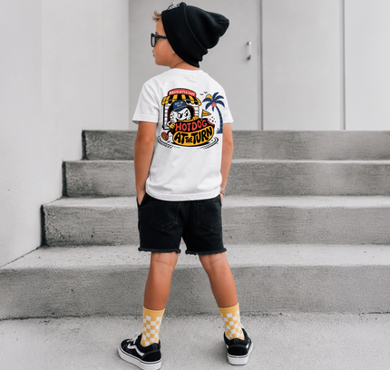 A child standing on concrete steps wearing a white graphic T-shirt, black beanie, black shorts, and yellow-and-white checkered socks. The shirt has a large colorful back graphic with the words 'HOTDOG ARTIST'.