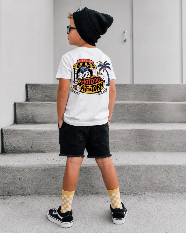 A child standing on concrete steps wearing a white graphic T-shirt, black beanie, black shorts, and yellow-and-white checkered socks. The shirt has a large colorful back graphic with the words 'HOTDOG ARTIST'.