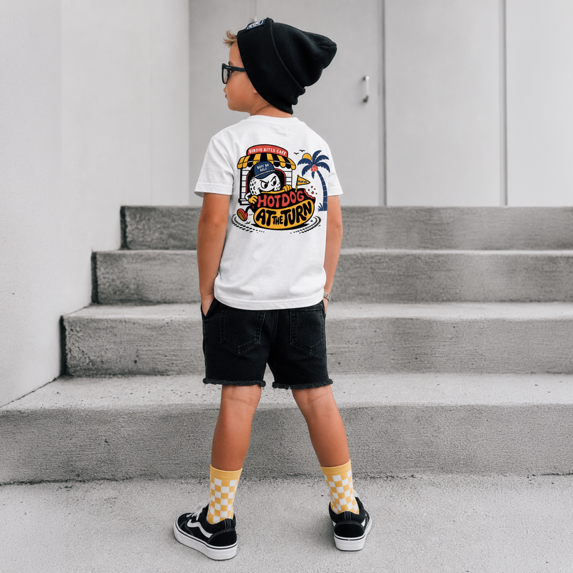 A child standing on concrete steps wearing a white graphic T-shirt, black beanie, black shorts, and yellow-and-white checkered socks. The shirt has a large colorful back graphic with the words 'HOTDOG ARTIST'.