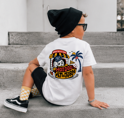 A child wearing a white graphic t-shirt with a 'Hotdog at the Jury' cartoon design on the back, sitting on concrete steps. The child is also wearing a black beanie, black sunglasses, black pants, and black-and-white checkered socks.