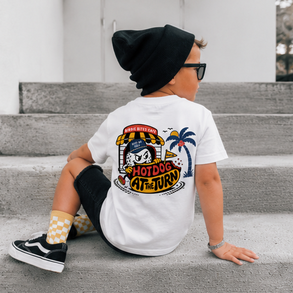 A child wearing a white graphic t-shirt with a 'Hotdog at the Jury' cartoon design on the back, sitting on concrete steps. The child is also wearing a black beanie, black sunglasses, black pants, and black-and-white checkered socks.
