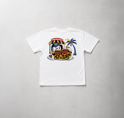 A white short-sleeve t-shirt laid flat on a light gray textured background, featuring a colorful retro-style graphic on the front with a cartoon character, palm tree, and the text 'HOTDOG AT THE END'.