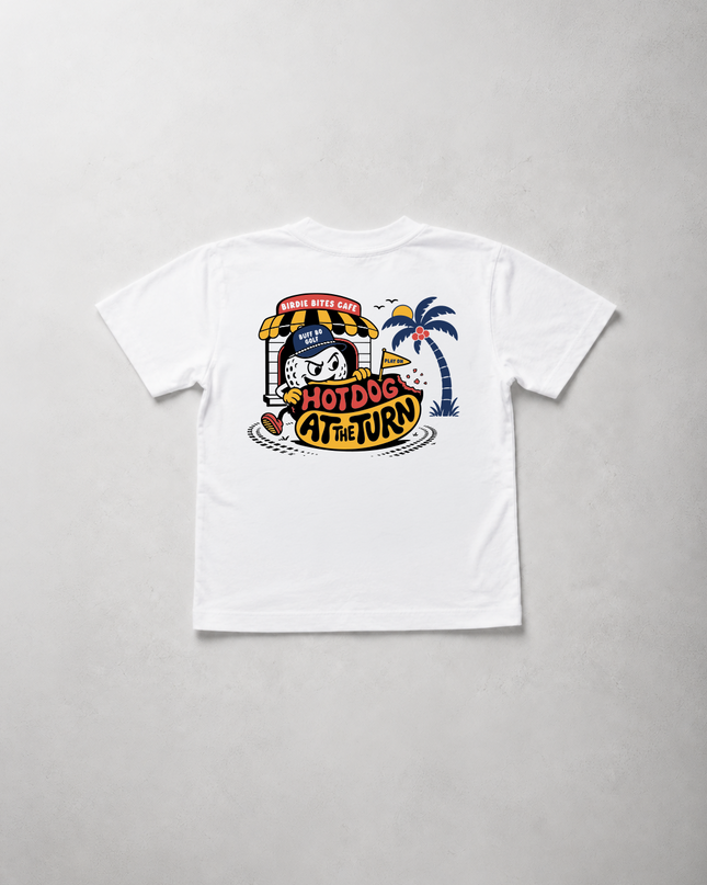 A white short-sleeve t-shirt laid flat on a light gray textured background, featuring a colorful retro-style graphic on the front with a cartoon character, palm tree, and the text 'HOTDOG AT THE END'.