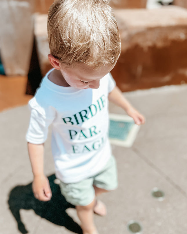 Young boy with blond hair wearing a white t-shirt that says "BIRDIE PAR EAGLE" in green text and light green shorts, walking barefoot on a concrete splash pad with embedded water jets; the background includes brown stone structures and bright sunlight casting distinct shadows.
