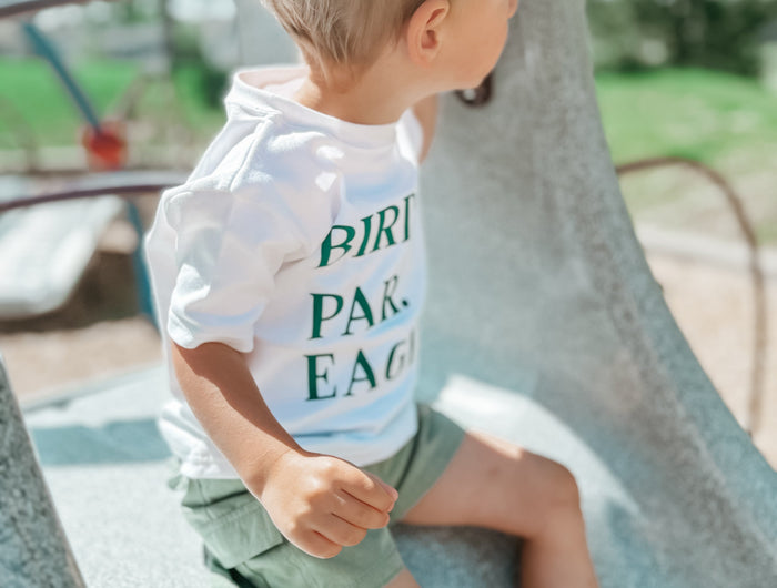 Young child wearing a white "BIRDIE. PAR. EAGLE." t-shirt and green shorts sits on a playground structure, looking to the side; the background features blurred trees, blue playground bars, and bright natural sunlight creating a soft, summery atmosphere.
