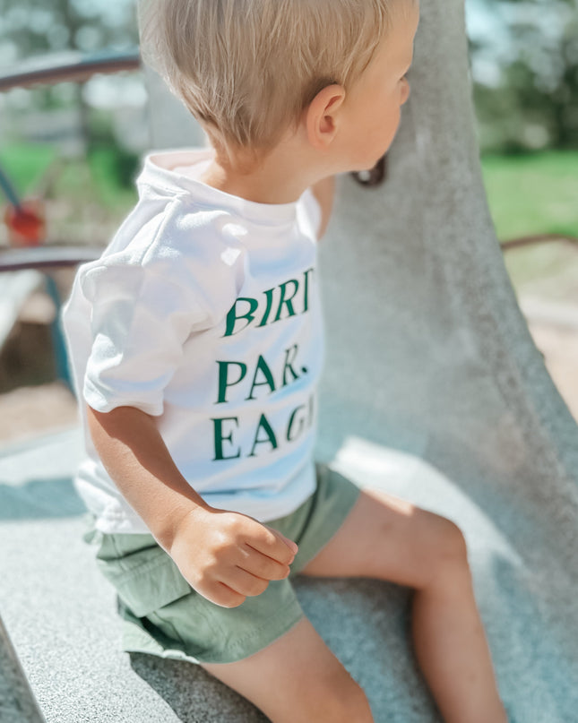 Young child wearing a white "BIRDIE. PAR. EAGLE." t-shirt and green shorts sits on a playground structure, looking to the side; the background features blurred trees, blue playground bars, and bright natural sunlight creating a soft, summery atmosphere.
