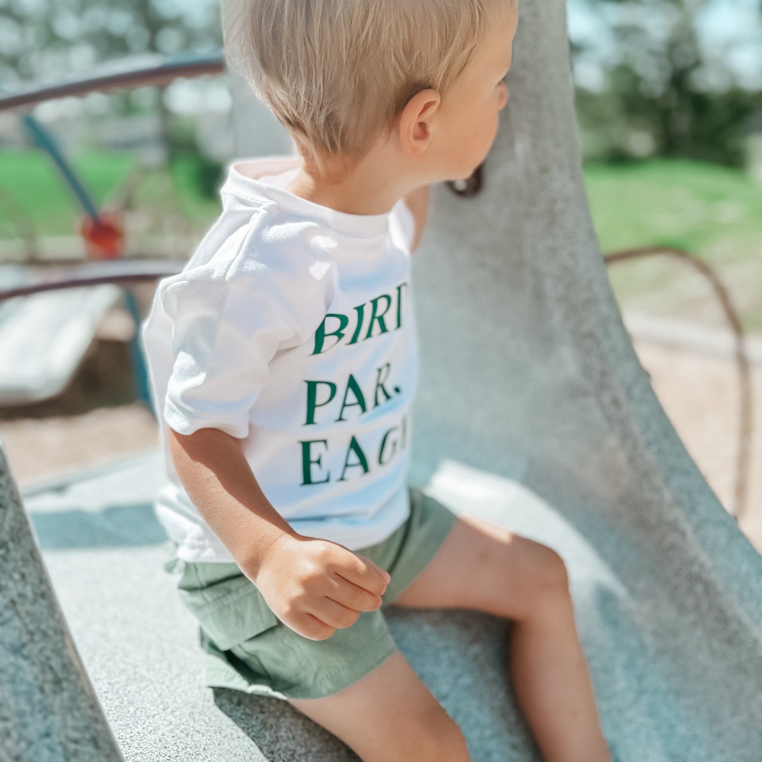 Young child wearing a white "BIRDIE. PAR. EAGLE." t-shirt and green shorts sits on a playground structure, looking to the side; the background features blurred trees, blue playground bars, and bright natural sunlight creating a soft, summery atmosphere.
