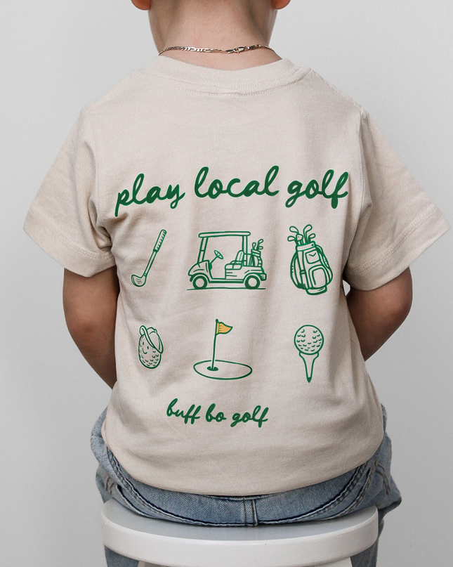 Child wearing a beige t-shirt with green golf-themed graphics and text, standing on a white stool against a light gray background.