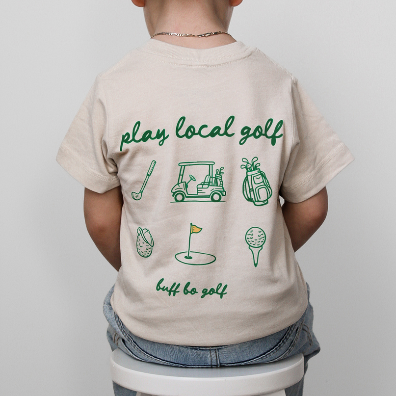 Child wearing a beige t-shirt with green golf-themed graphics and text, standing on a white stool against a light gray background.