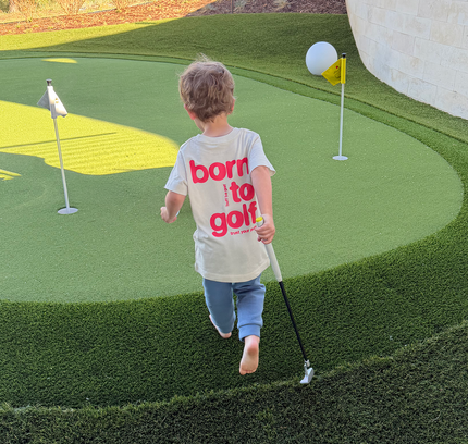 Child playing golf on a putting green with a 'born to golf' shirt.