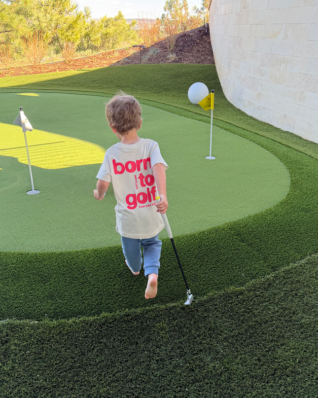Child playing golf on a putting green with a 'born to golf' shirt.