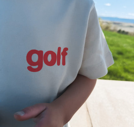 Beige t-shirt with 'golf' in red text worn by a person outdoors.