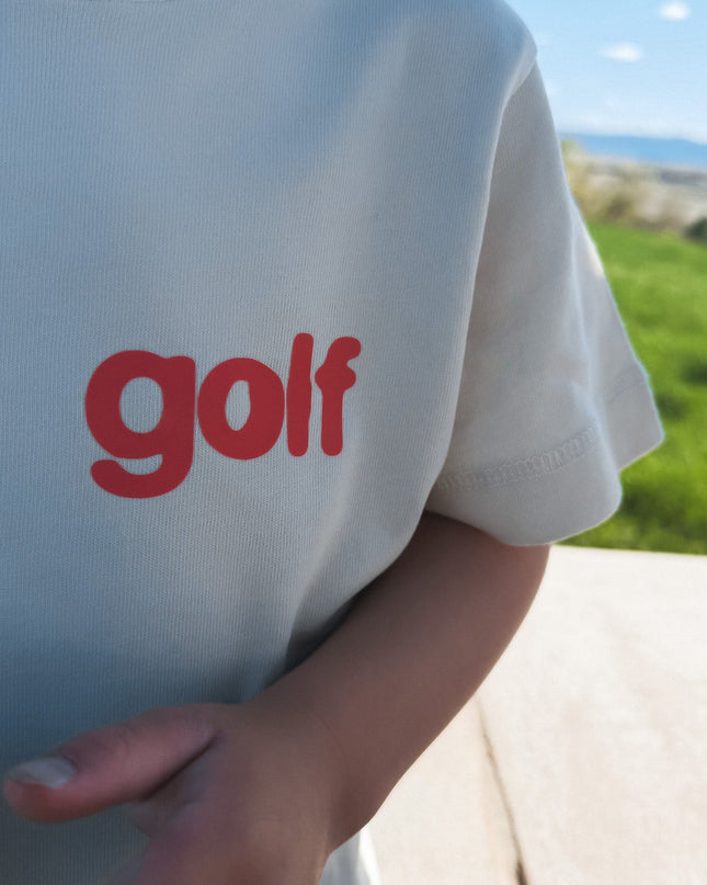 Beige t-shirt with 'golf' in red text worn by a person outdoors.