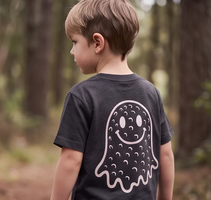 A young boy seen from behind wearing a dark charcoal gray t-shirt with a large white ghost graphic printed on the back, standing in a blurred forest setting.