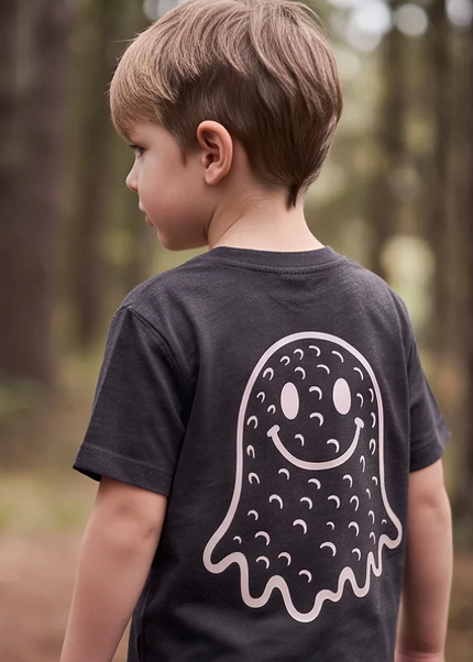 A young boy seen from behind wearing a dark charcoal gray t-shirt with a large white ghost graphic printed on the back, standing in a blurred forest setting.