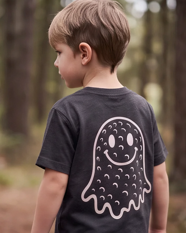 A young boy seen from behind wearing a dark charcoal gray t-shirt with a large white ghost graphic printed on the back, standing in a blurred forest setting.