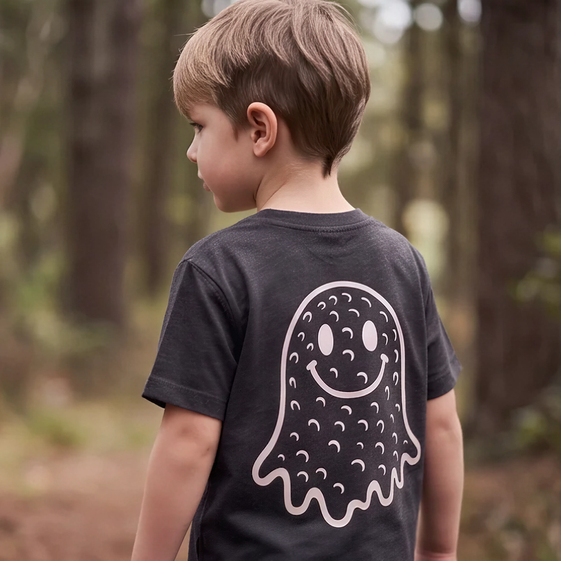 A young boy seen from behind wearing a dark charcoal gray t-shirt with a large white ghost graphic printed on the back, standing in a blurred forest setting.