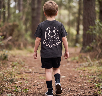 A young boy walking away on a forest path, wearing a dark charcoal gray t-shirt with a white ghost graphic on the back, black shorts, and black socks.