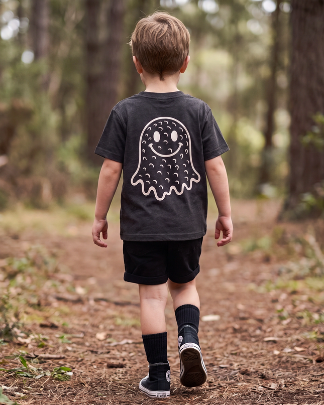 A young boy walking away on a forest path, wearing a dark charcoal gray t-shirt with a white ghost graphic on the back, black shorts, and black socks.