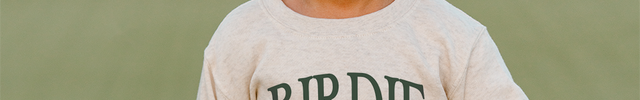Child wearing a cap and sweatshirt with 'NERDY FORE' and 'BIRDIE' text, standing on a golf course.