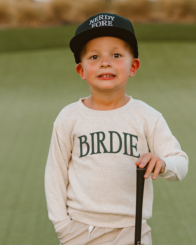 Child wearing a cap and sweatshirt with 'NERDY FORE' and 'BIRDIE' text, standing on a golf course.