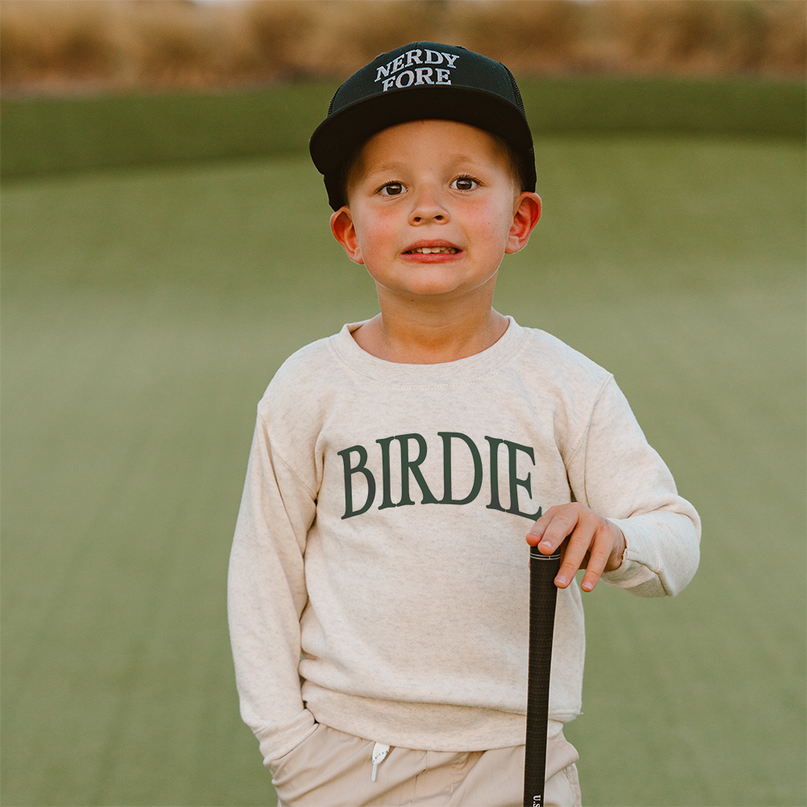 Child wearing a cap and sweatshirt with 'NERDY FORE' and 'BIRDIE' text, standing on a golf course.