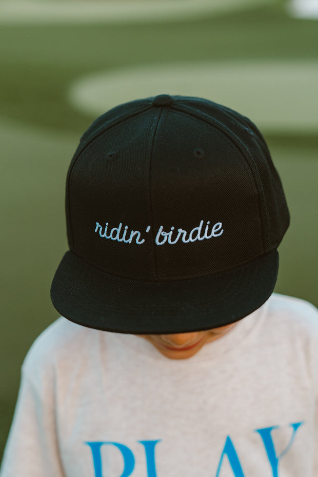 Black cap with 'ridin' birdie' text worn by a kid, blurred background