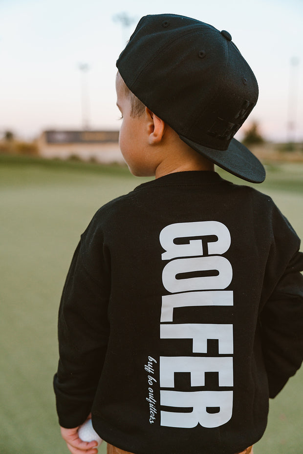 Child wearing a black long-sleeve shirt with 'GOLFER' printed on the back, standing on a golf course.