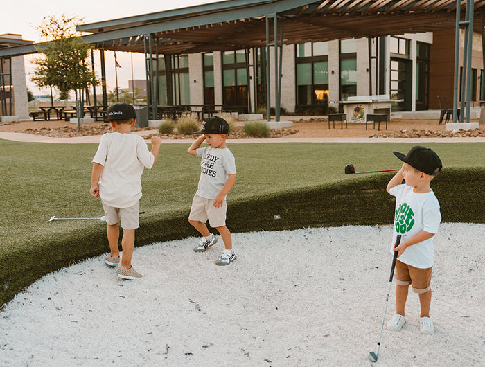Three children playing golf on a putting green with a building in the background.