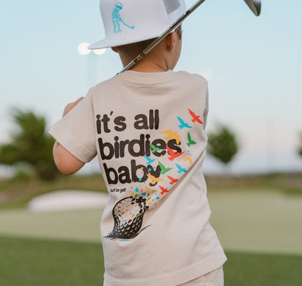Child holding a golf club on a golf course wearing a shirt with a humorous golf-themed design.
