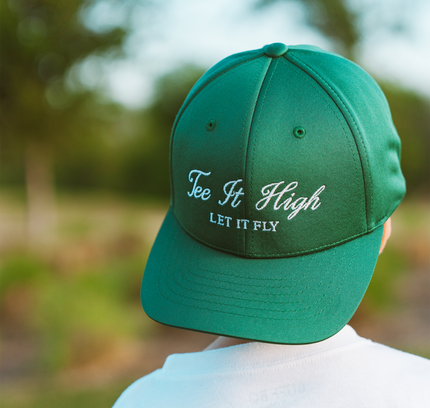 Green cap with 'Tee It High, Let It Fly' text worn by a person in a golf-themed shirt outdoors.