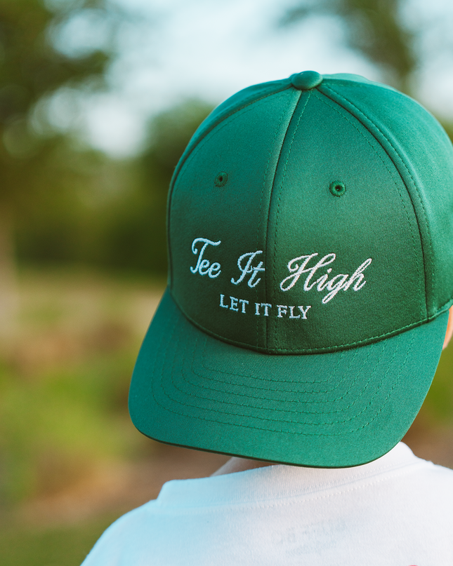 Green cap with 'Tee It High, Let It Fly' text worn by a person in a golf-themed shirt outdoors.