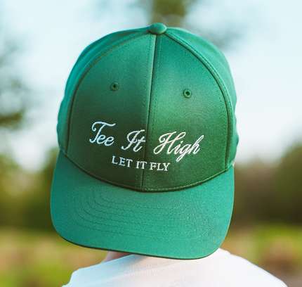 Green cap with 'Tee It High Let It Fly' text against a blurred outdoor background