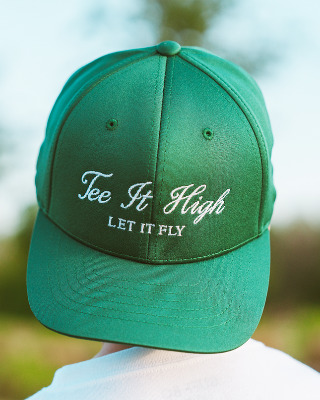 Green cap with 'Tee It High Let It Fly' text against a blurred outdoor background
