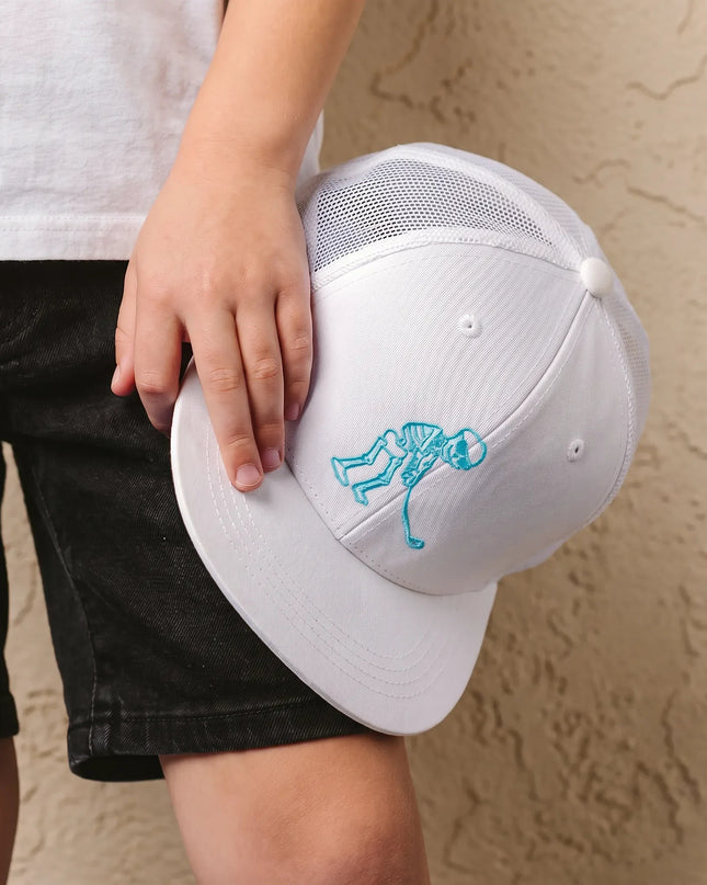 A child holding a white baseball cap with a light blue embroidered skeleton design, wearing a white t-shirt and black denim shorts against a textured beige wall.