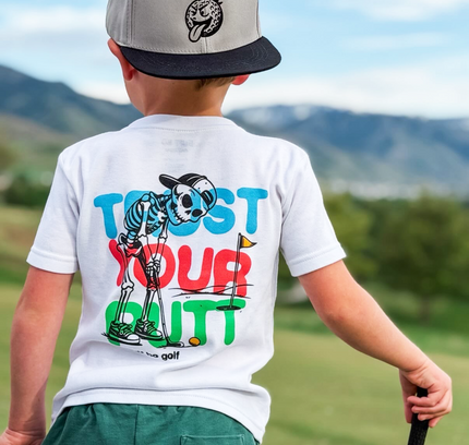 Child wearing a white t-shirt with colorful graphics and text, standing on a golf course.