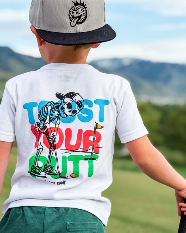 Child wearing a white t-shirt with colorful graphics and text, standing on a golf course.
