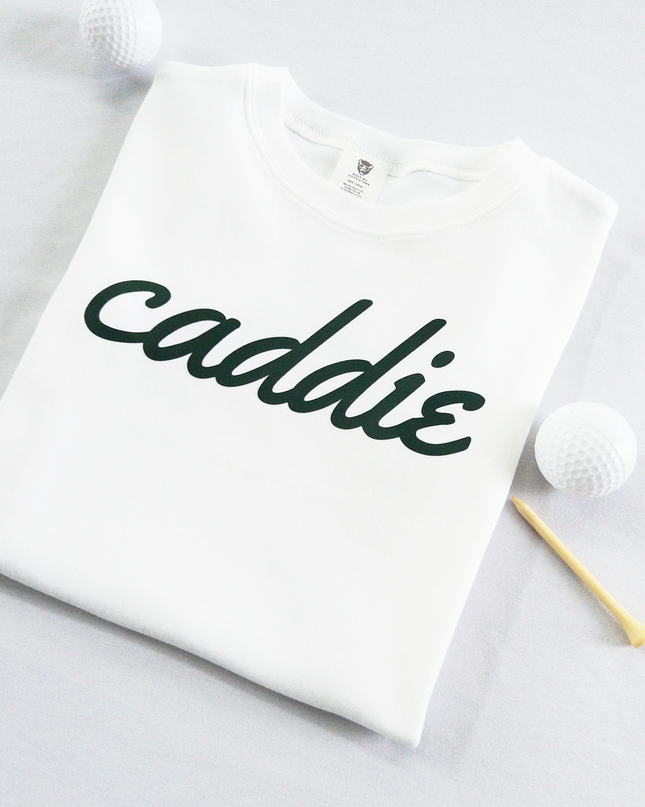 White kids golf t-shirt with dark green cursive text reading 'caddie,' displayed flat with golf balls and a wooden tee, perfect for young golf enthusiasts and future caddies.