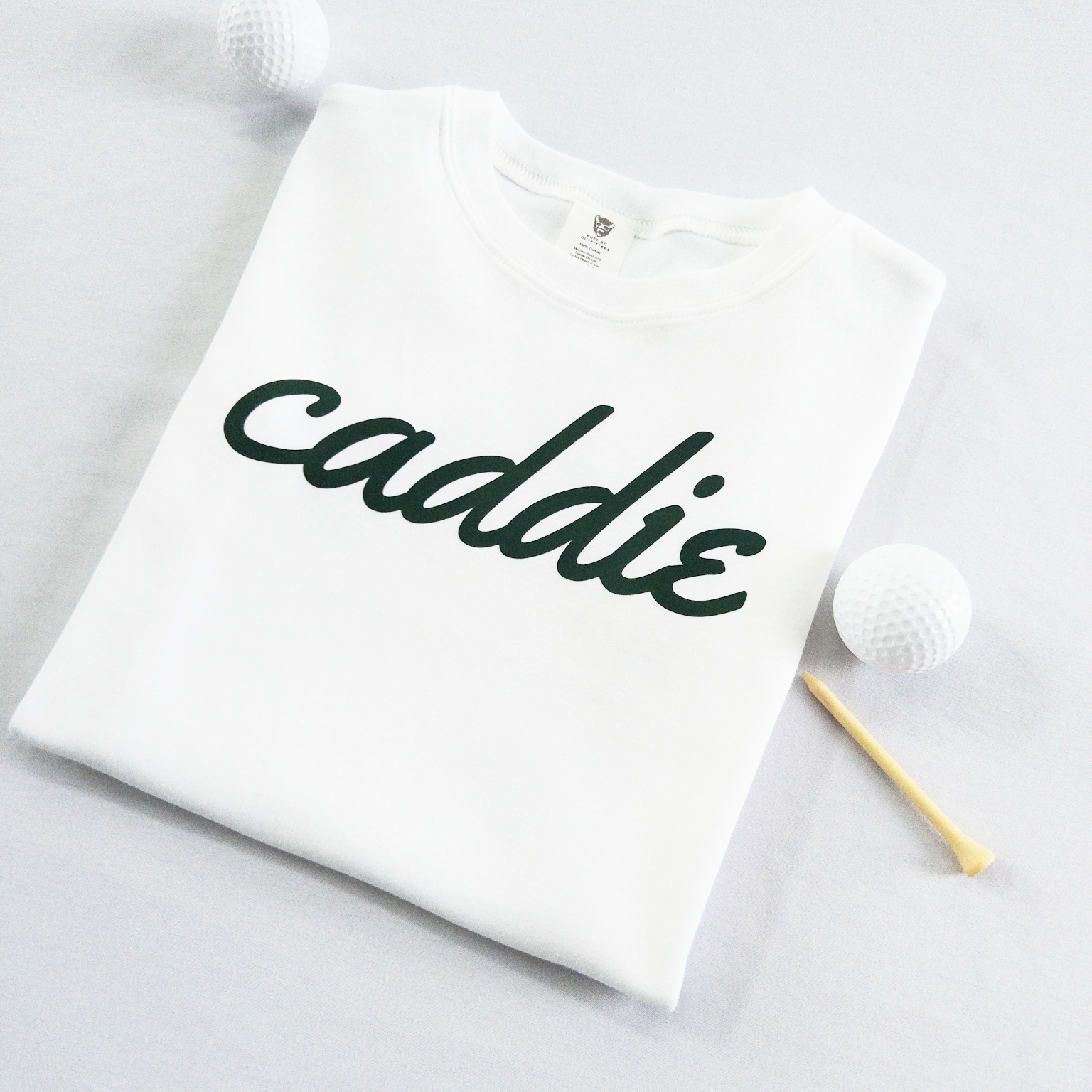 White kids golf t-shirt with dark green cursive text reading 'caddie,' displayed flat with golf balls and a wooden tee, perfect for young golf enthusiasts and future caddies.