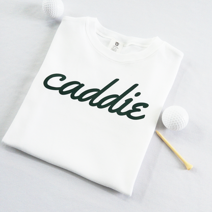 White kids golf t-shirt with dark green cursive text reading 'caddie,' displayed flat with golf balls and a wooden tee, perfect for young golf enthusiasts and future caddies.